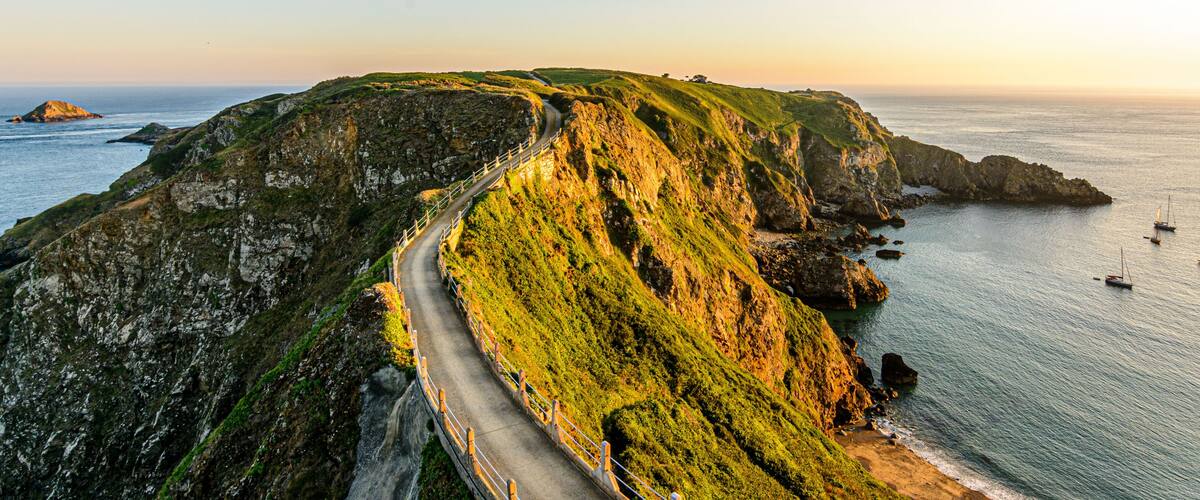 A coastal road winding along a cliffside, overlooking the sea with boats in the distance.