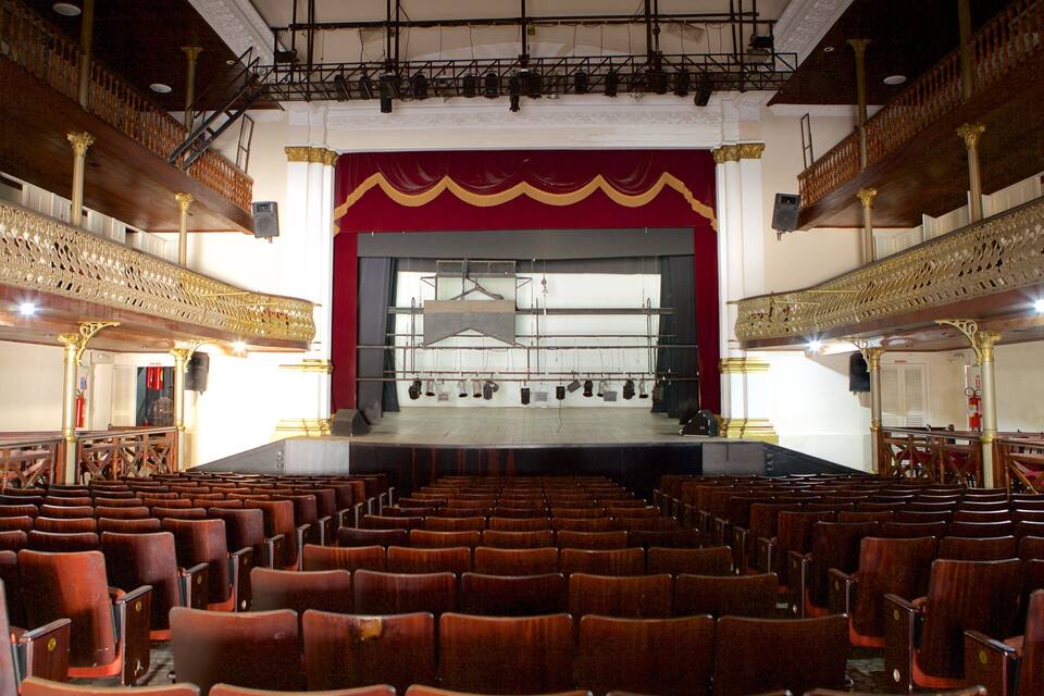 Um teatro vazio com grades douradas ornamentadas e um palco com fundo vermelho.