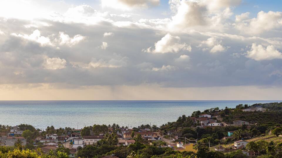 A coastal town with houses, palm trees, and a view of the sea under a partly cloudy sky.