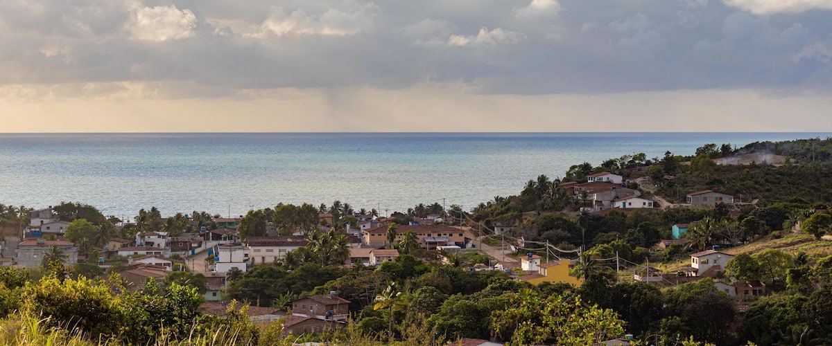 A coastal town with houses, palm trees, and a view of the sea under a partly cloudy sky.
