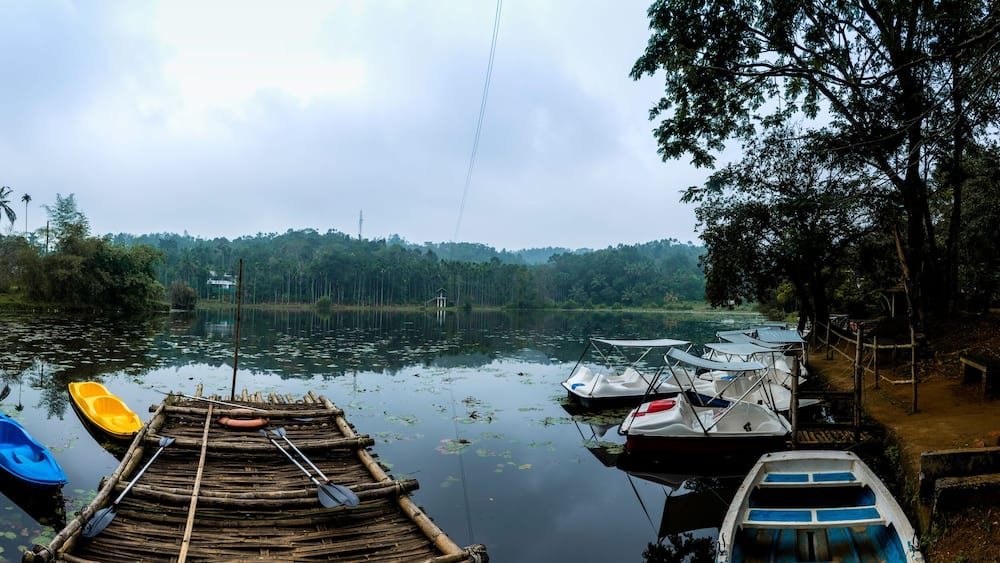 Un lac paisible, avec des bateaux amarrés sur la rive, entouré d’une forêt dense et surplombé d’un ciel nuageux.