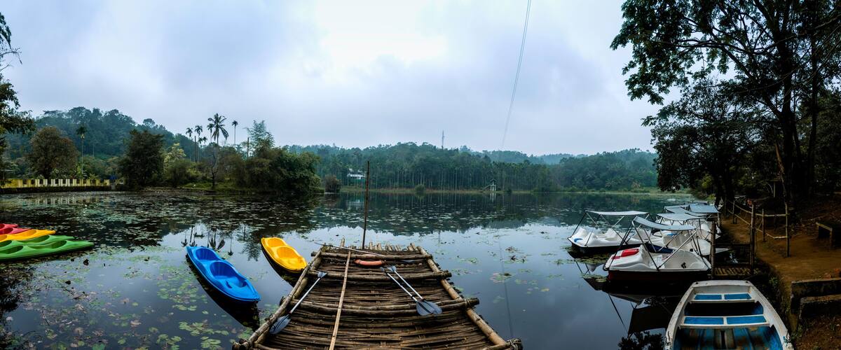 A calm lake with boats docked on the shore, surrounded by dense trees and a cloudy sky.