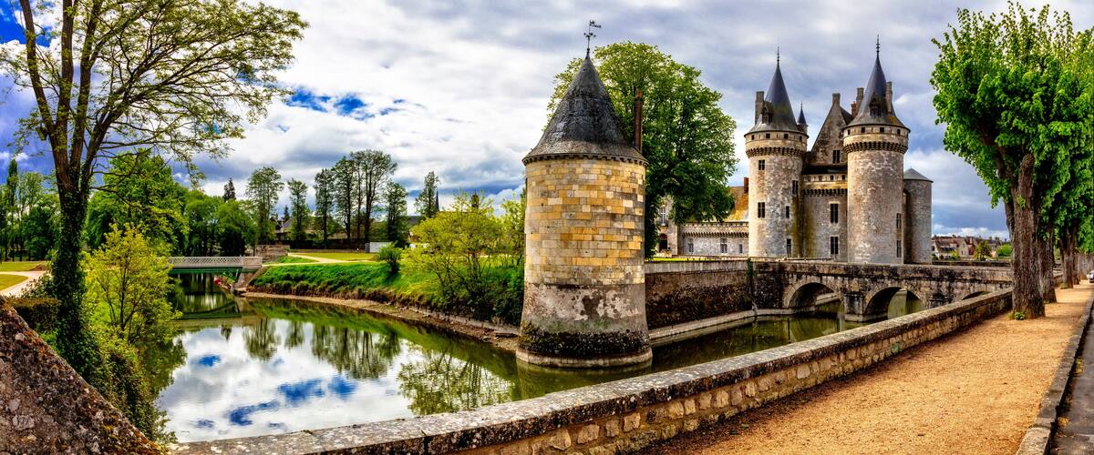 A historic castle with multiple towers, a moat, and lush greenery.