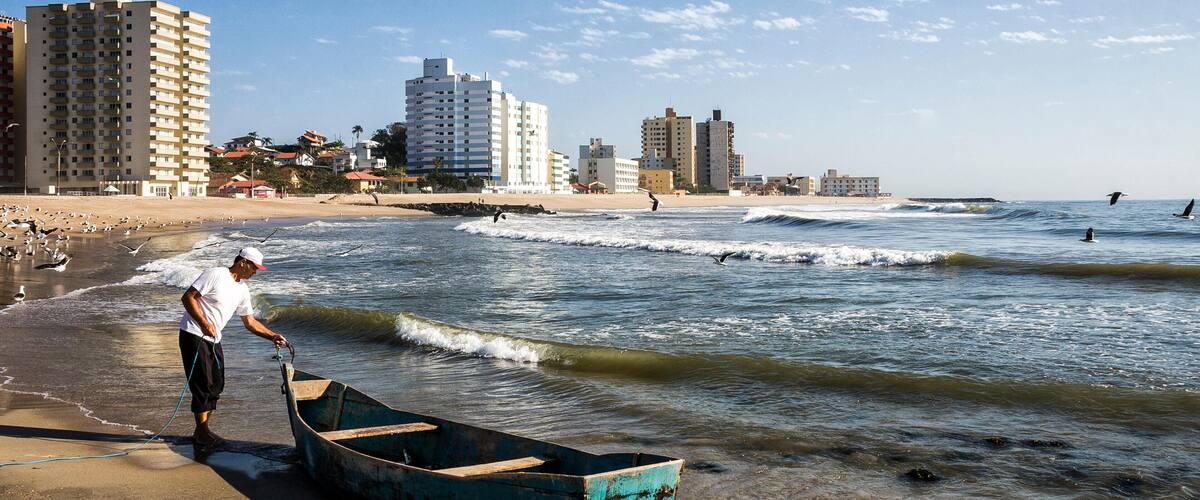 A beach with a man pulling a boat onto the shore, waves in the background, and buildings in the distance.