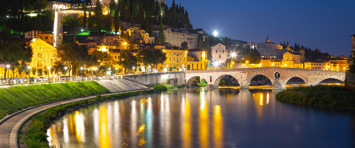 A river with a stone bridge, illuminated buildings, and a castle on a hill.