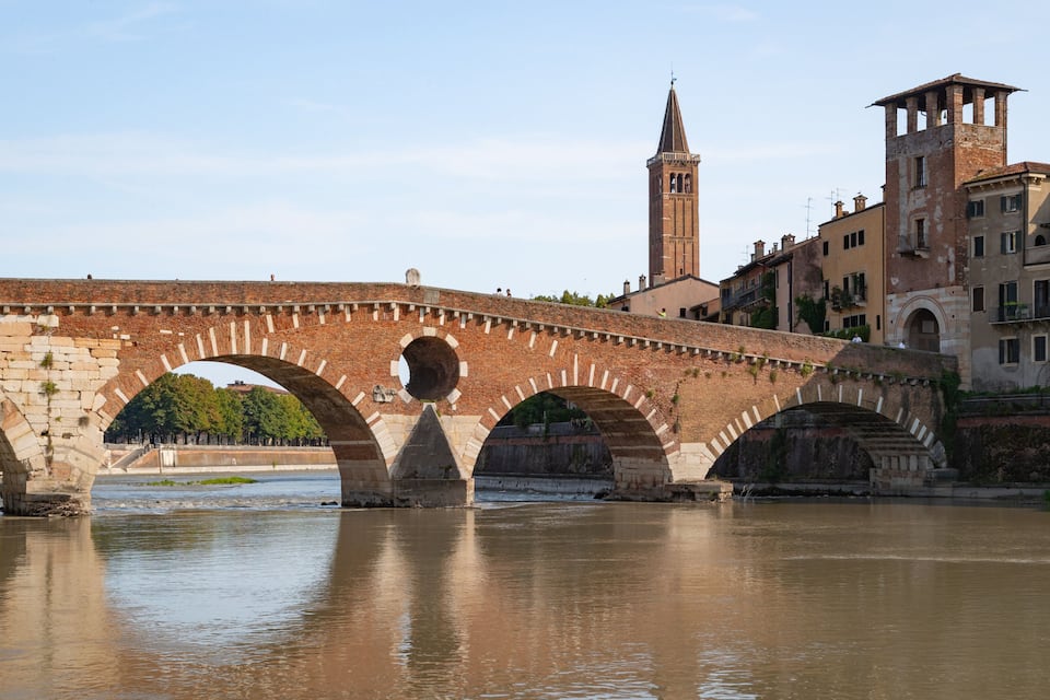 A stone bridge with multiple arches over a river, a tower, and buildings in the background.