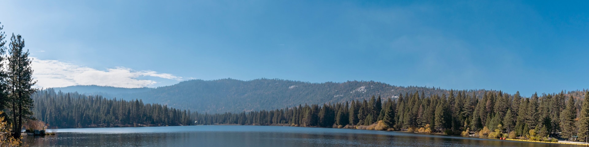 Un lago tranquilo rodeado de bosques densos y montañas lejanas bajo un cielo despejado.