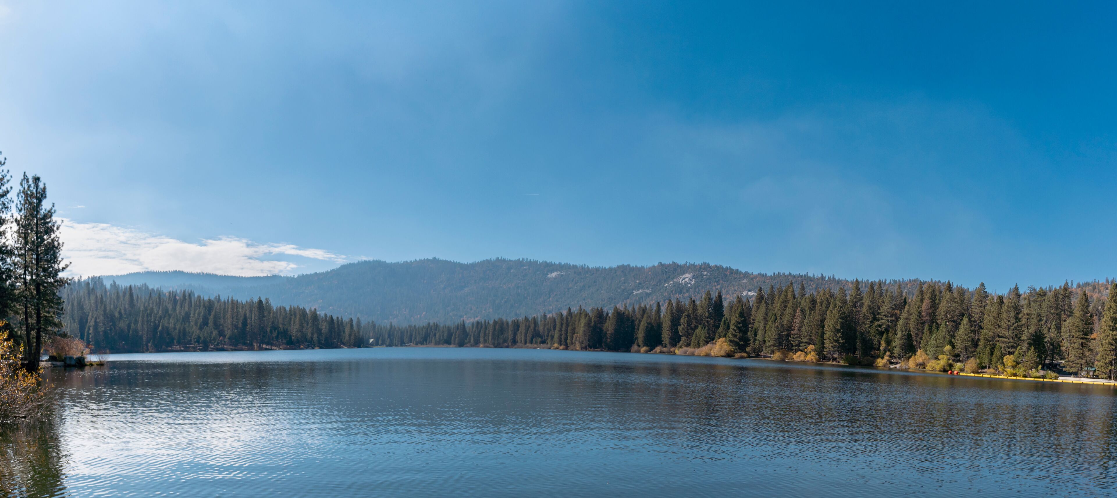 A calm lake surrounded by dense forests and distant mountains under a clear sky.