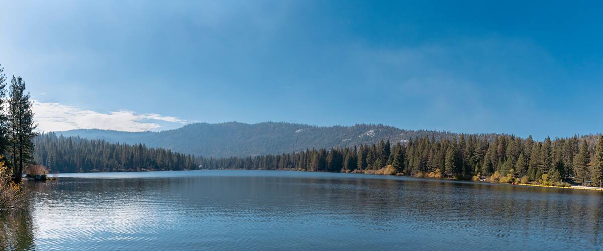 A calm lake surrounded by dense forests and distant mountains under a clear sky.