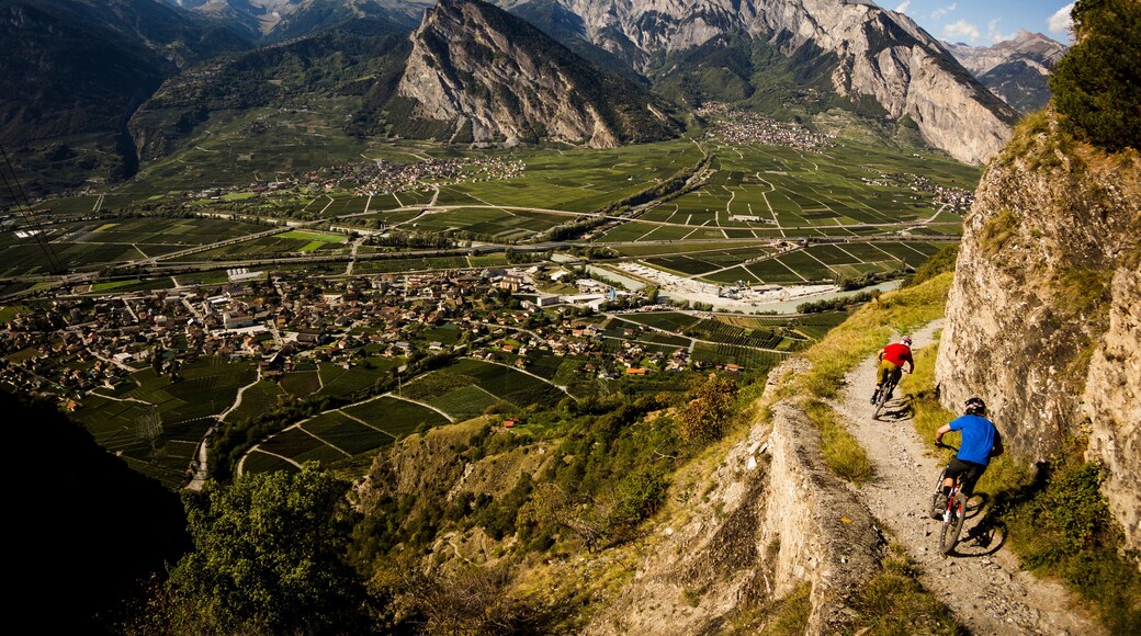 RIDDES, SWITZERLAND. Two men ride mountain bikes along a narrow exposed trail in the Swiss Alps. The dramatic cliff in the background is the Haut de Cry.