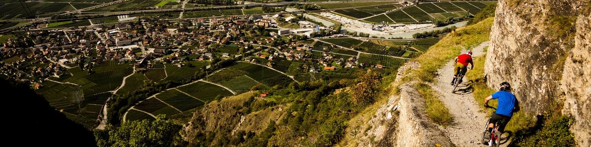 RIDDES, SWITZERLAND. Two men ride mountain bikes along a narrow exposed trail in the Swiss Alps. The dramatic cliff in the background is the Haut de Cry.