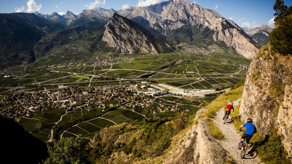 RIDDES, SWITZERLAND. Two men ride mountain bikes along a narrow exposed trail in the Swiss Alps. The dramatic cliff in the background is the Haut de Cry.