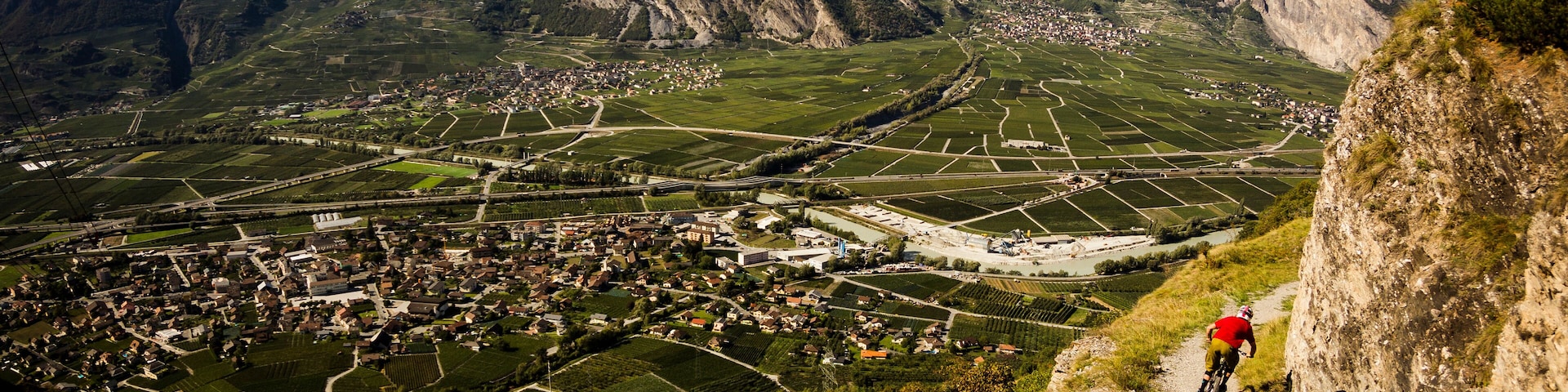 RIDDES, SWITZERLAND. Two men ride mountain bikes along a narrow exposed trail in the Swiss Alps. The dramatic cliff in the background is the Haut de Cry.