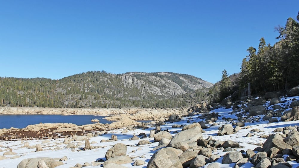 A snowy landscape with large rocks, a clear blue sky, and a forested mountain in the background.