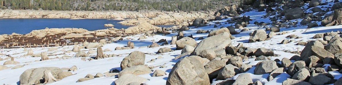A snowy landscape with large rocks, a clear blue sky, and a forested mountain in the background.
