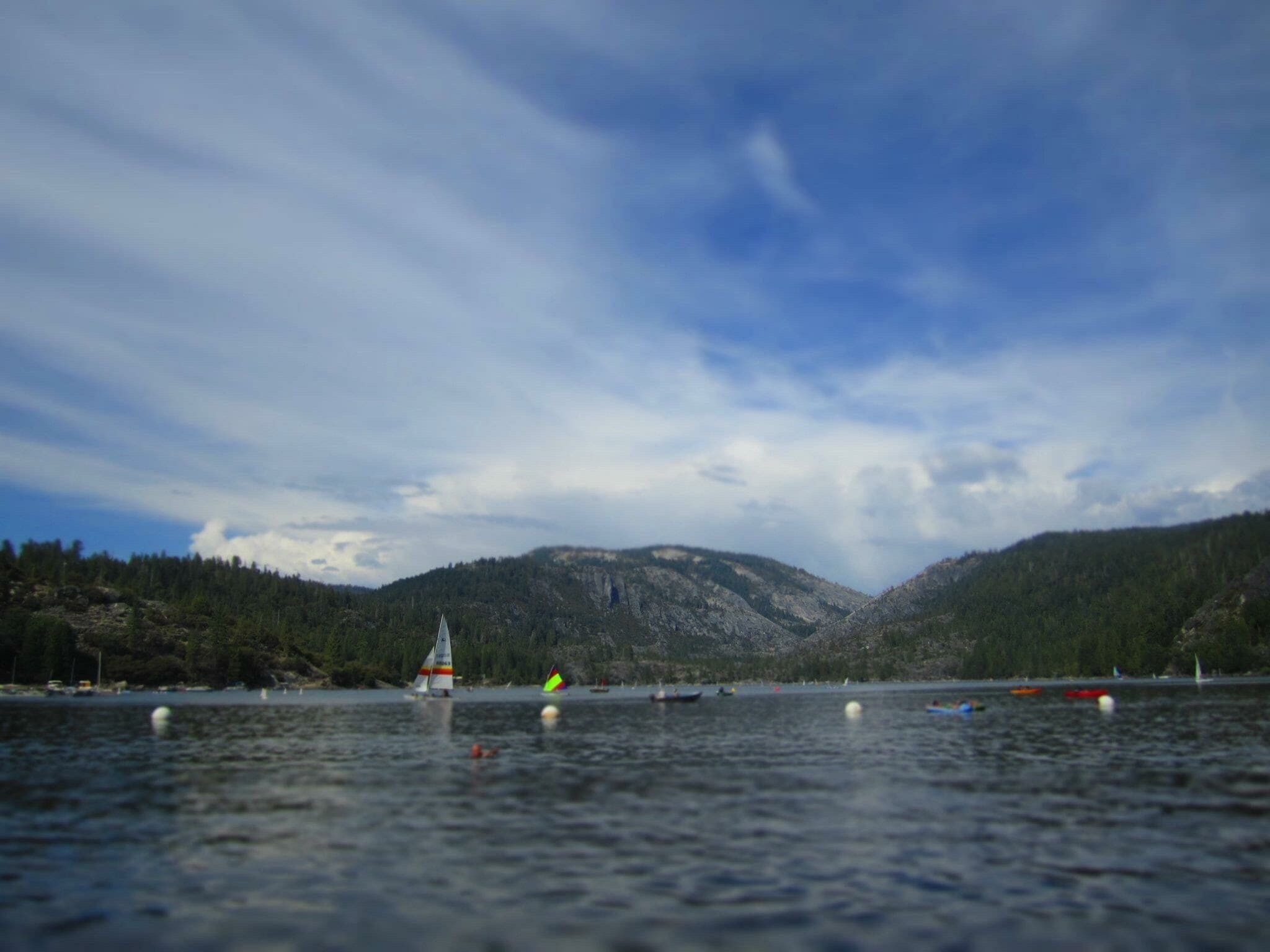 A calm lake with sailboats, surrounded by forested hills under a partly cloudy sky.