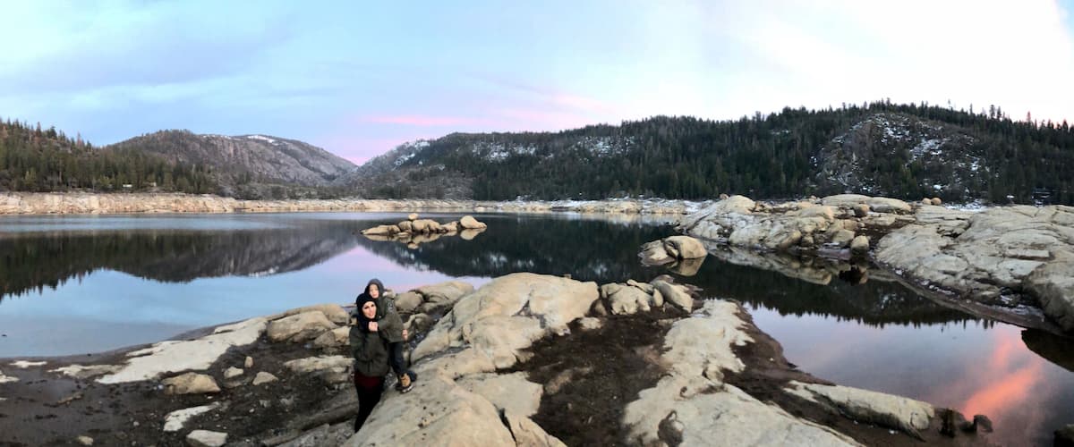 A calm lake with rocky shores, surrounded by forested mountains and a clear sky.