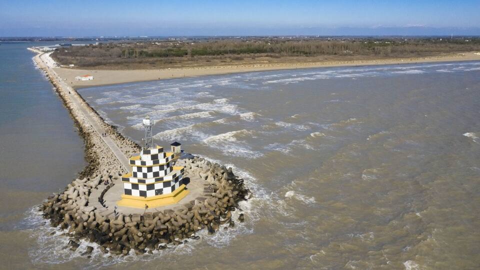 A coastal area with a checkered lighthouse on a rocky outcrop, a sandy beach, and a distant mountain range.