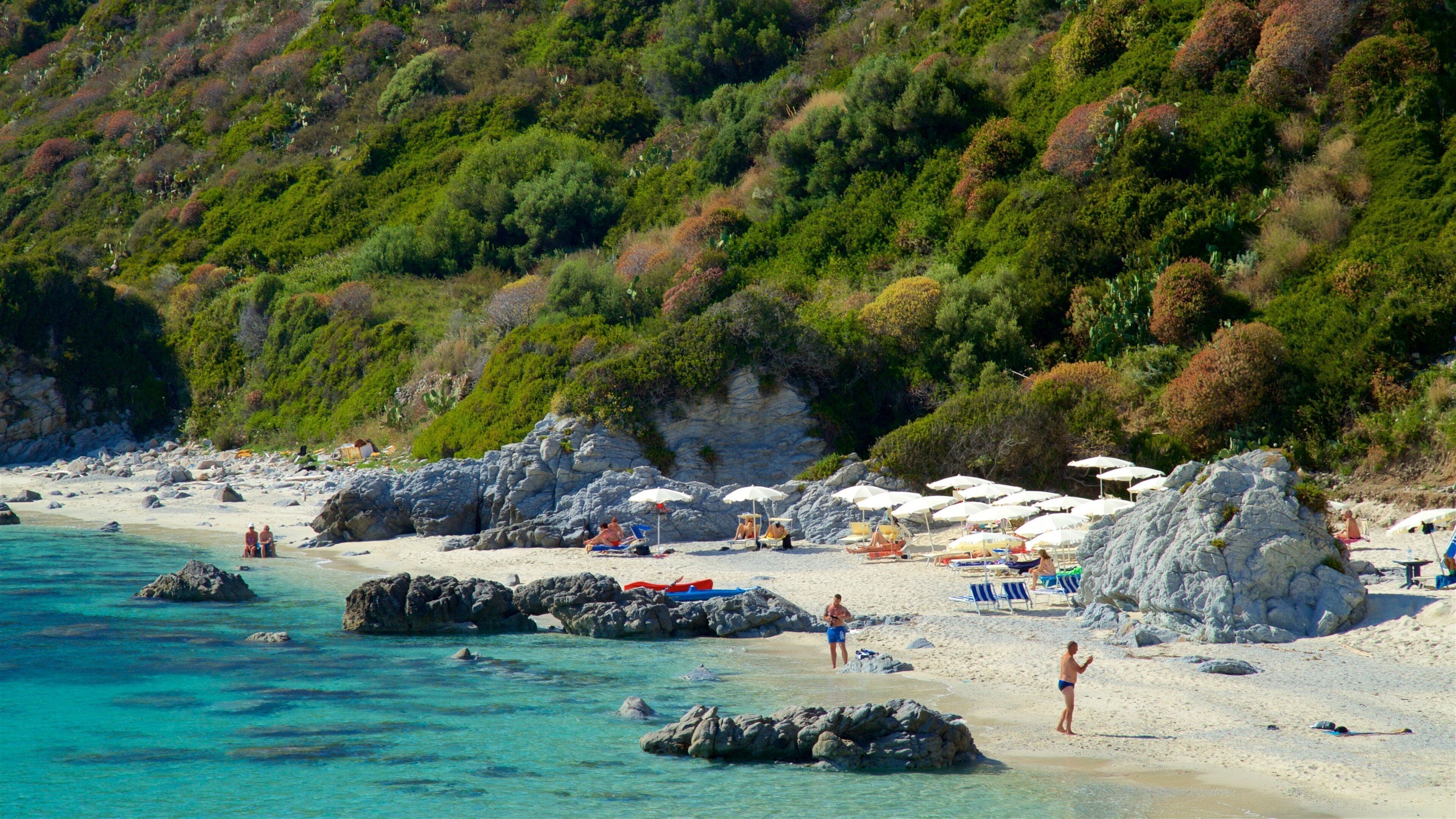 A beach with clear turquoise water, rocky outcrops, and lush green hills in the background.