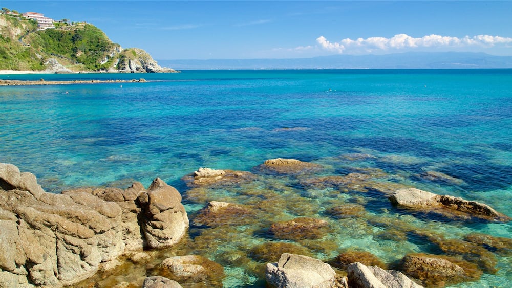 Un paysage côtier caractérisé par une eau turquoise et limpide, des formations rocheuses et une colline lointaine parsemée de bâtiments.