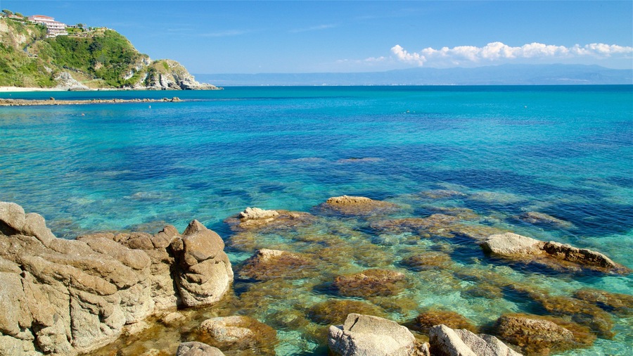 A coastal landscape with clear turquoise waters, rocky formations, and a distant hill with buildings.