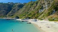 A coastal beach with clear turquoise waters, rocky outcrops, and a hillside with vegetation.