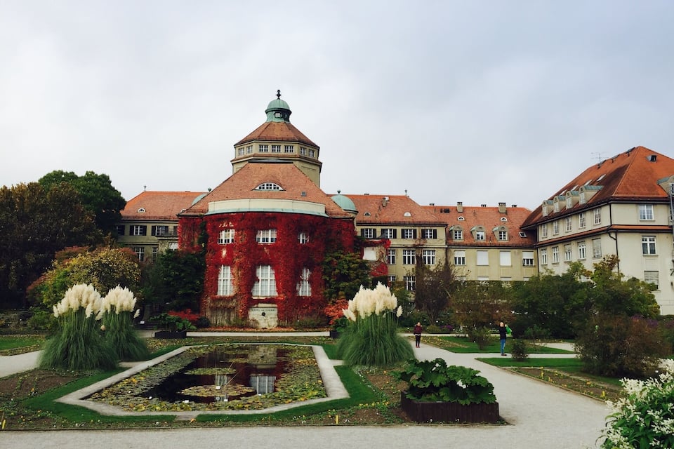 Ein historisches Gebäude mit roter Fassade und einem zentralen Turm, umgeben von einem formalen Garten mit Teich und gepflegten Rasenflächen.