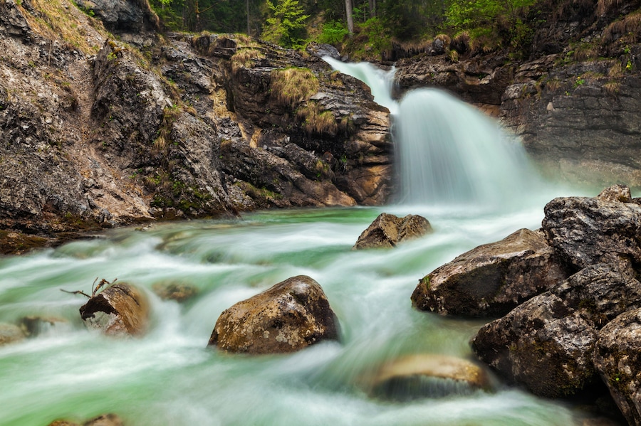 Une cascade dâeau qui dĂ©vale des rochers pour se jeter dans une riviĂšre, le tout entourĂ© de collines boisĂ©es.