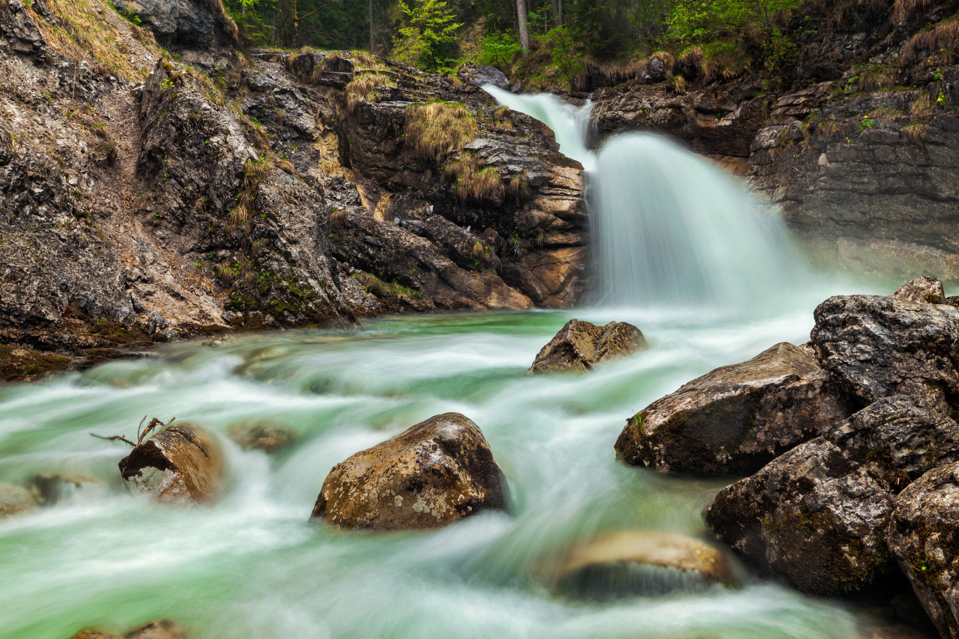 A waterfall cascading over rocks into a river, surrounded by forested hills.