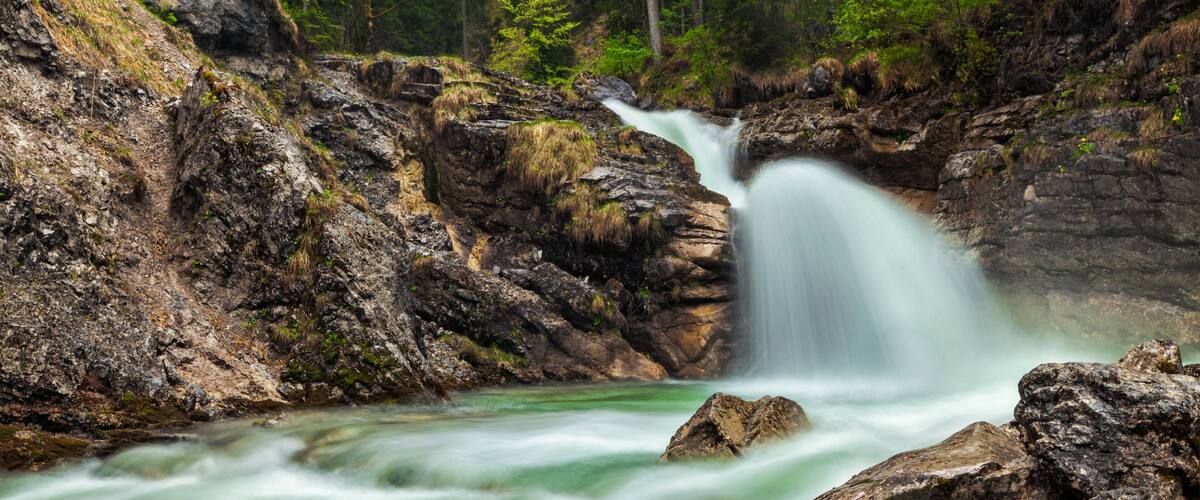 A waterfall cascading over rocks into a river, surrounded by forested hills.