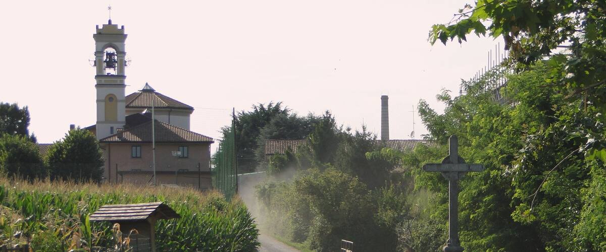 A rural road curving through a green landscape with a church tower, a small bridge, and a canal.