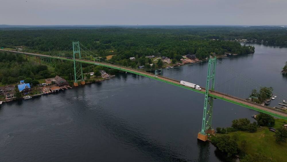 Un pont suspendu vert enjambant une rivière, entouré d’une forêt dense et de petits bâtiments.