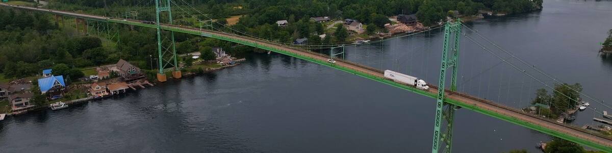 A green suspension bridge over a river, surrounded by dense forest and small buildings.