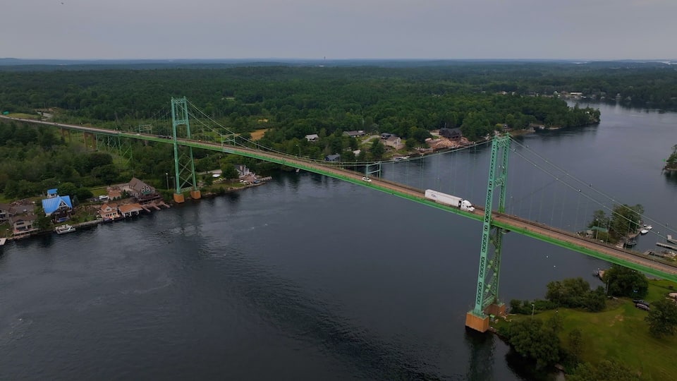 A green suspension bridge over a river, surrounded by dense forest and small buildings.