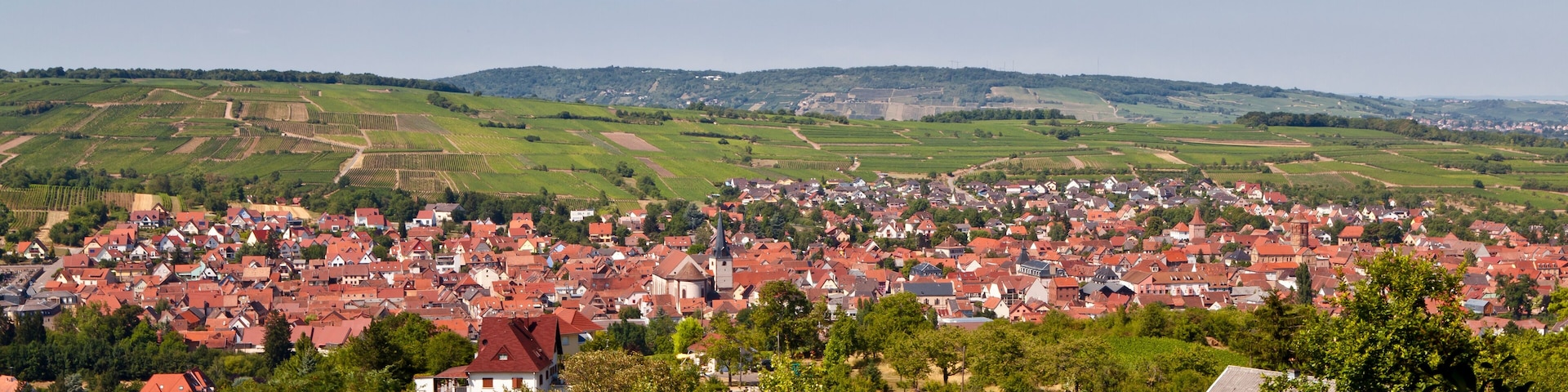 A town with red-roofed houses, green fields, and rolling hills in the background.