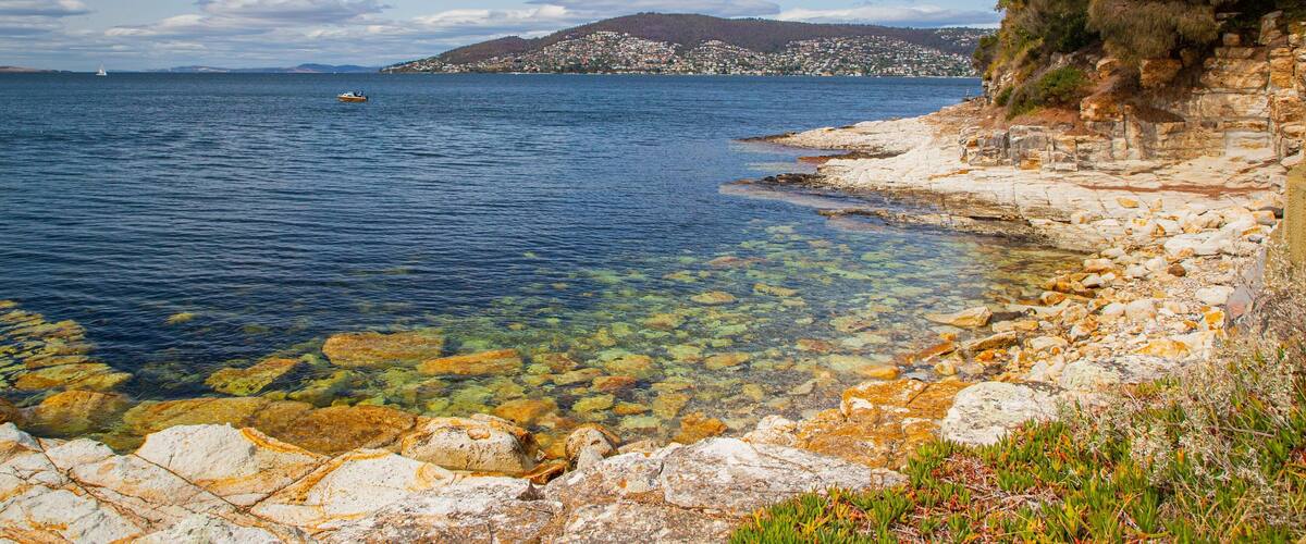 A coastal landscape with clear shallow waters revealing colorful rocks, a rocky shoreline, and a distant hill with buildings.