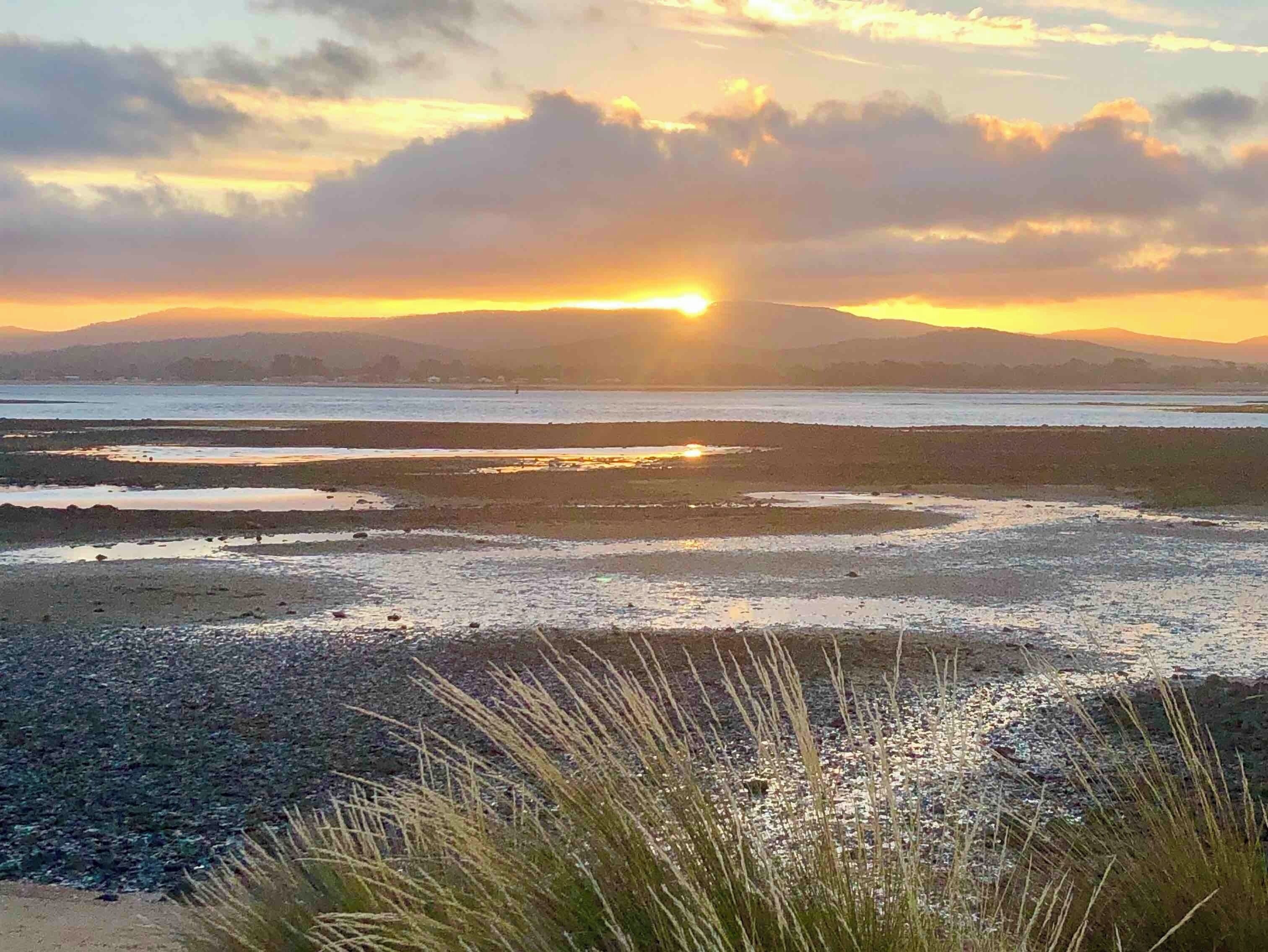 A sunset over a coastal landscape with a beach and distant mountains.