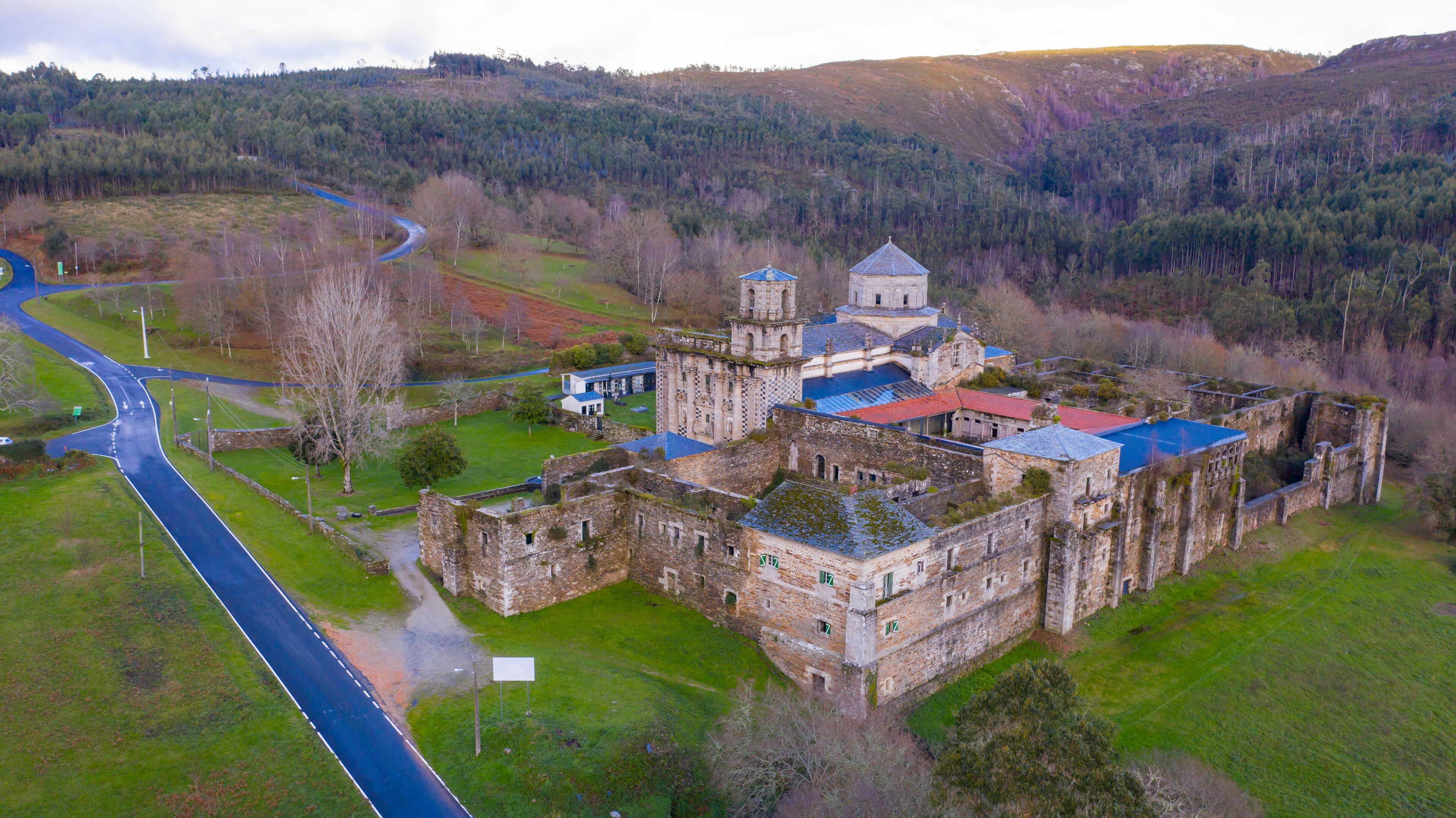 An aerial view of a historic castle complex with a red-roofed building, a tower, and surrounding green fields and hills.