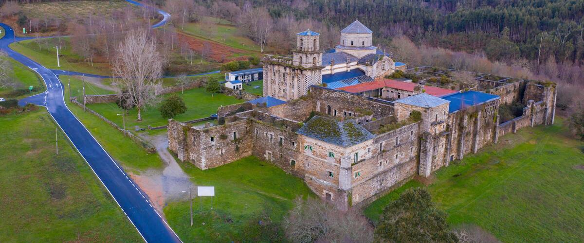 An aerial view of a historic castle complex with a red-roofed building, a tower, and surrounding green fields and hills.