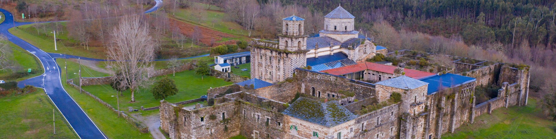 An aerial view of a historic castle complex with a red-roofed building, a tower, and surrounding green fields and hills.