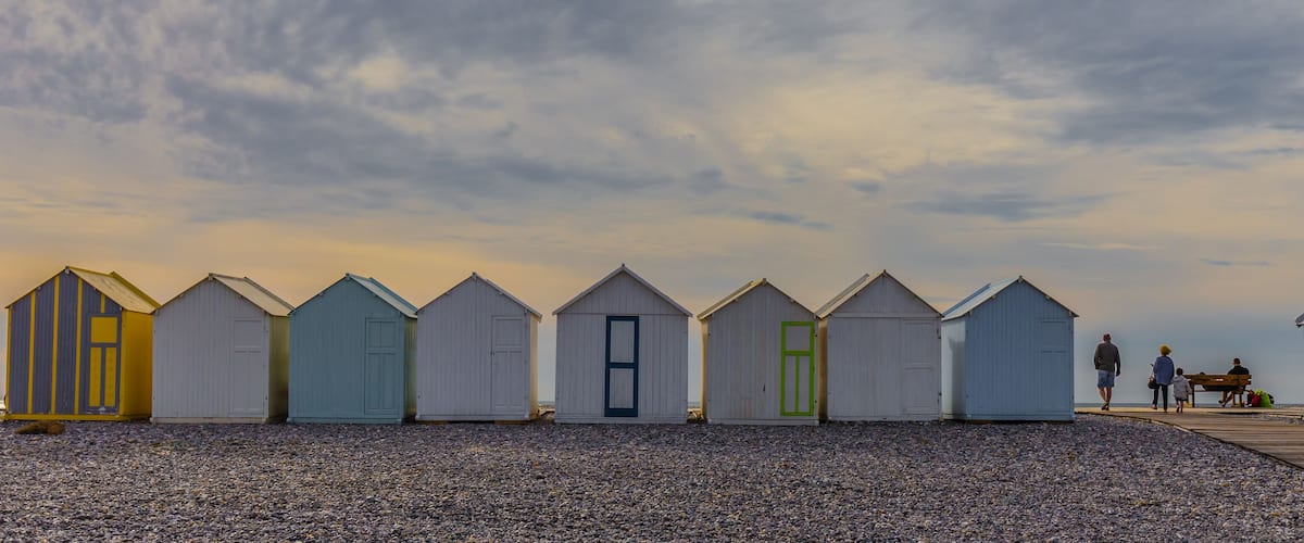 A row of beach huts on a pebbled beach with a wooden walkway leading to a bench area.