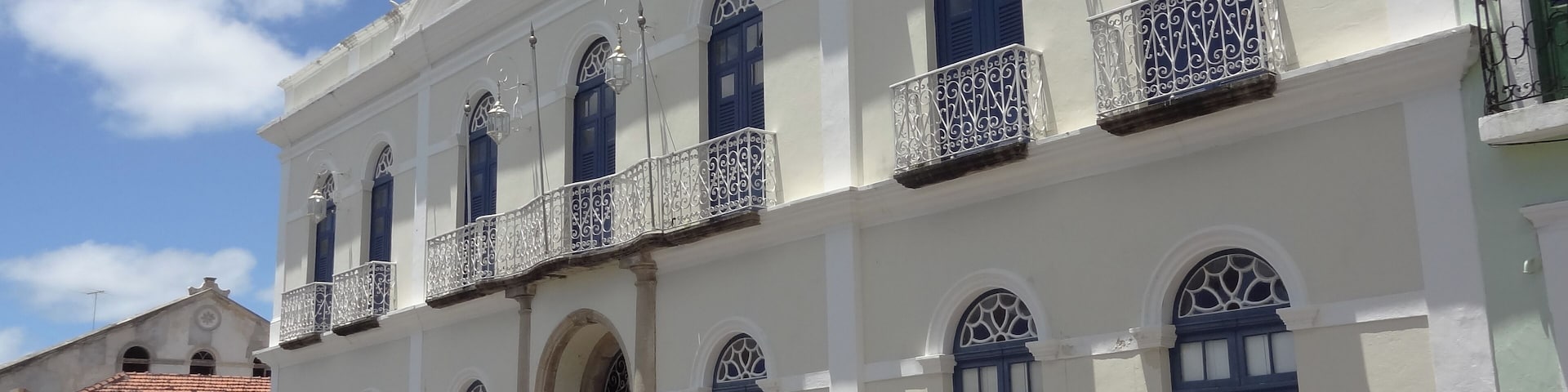 Um edifício em estilo colonial com paredes brancas e persianas azuis, com varandas ornamentadas e janelas em arco.
