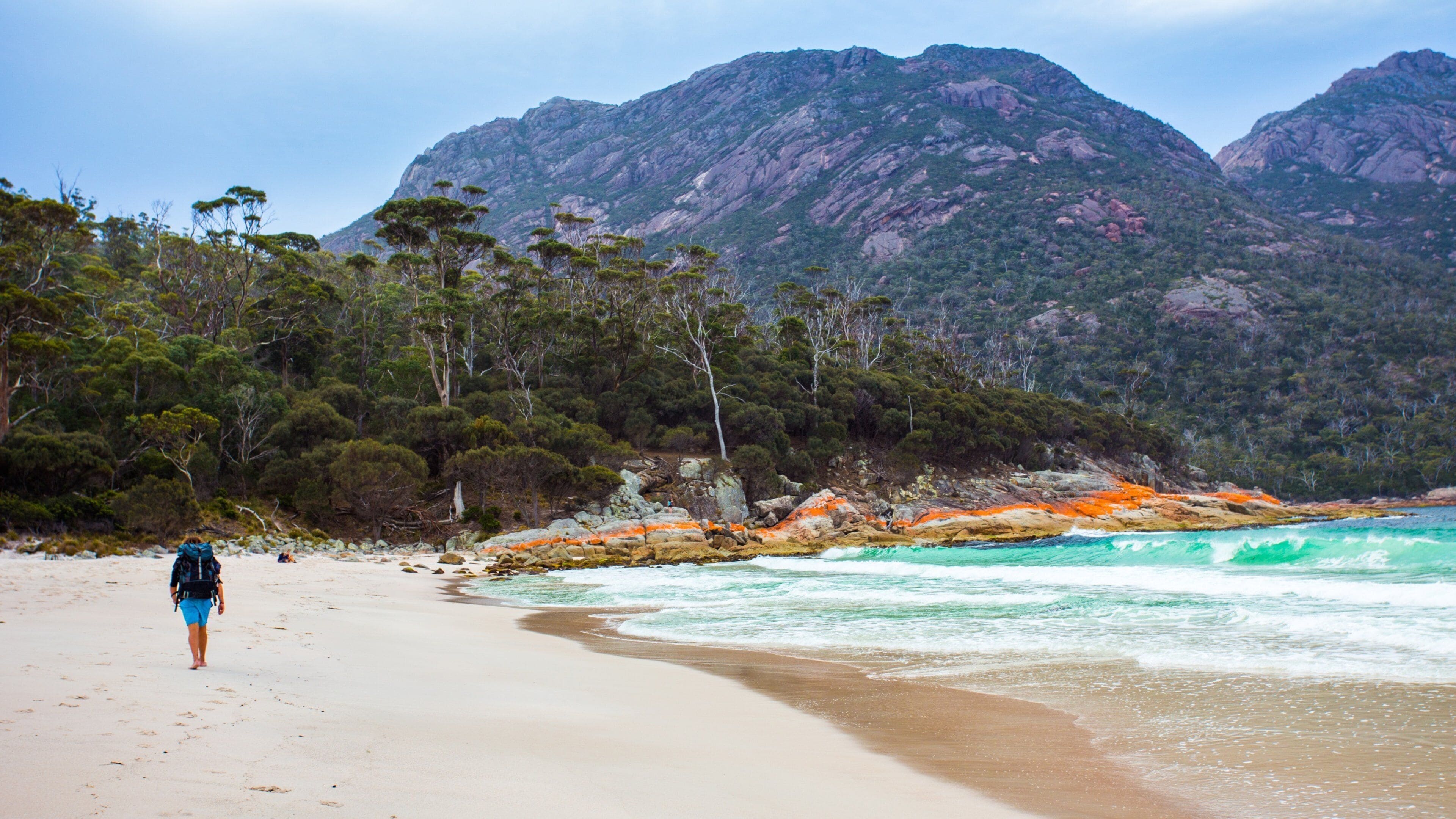 A beach with a person walking, surrounded by rocky outcrops and trees.