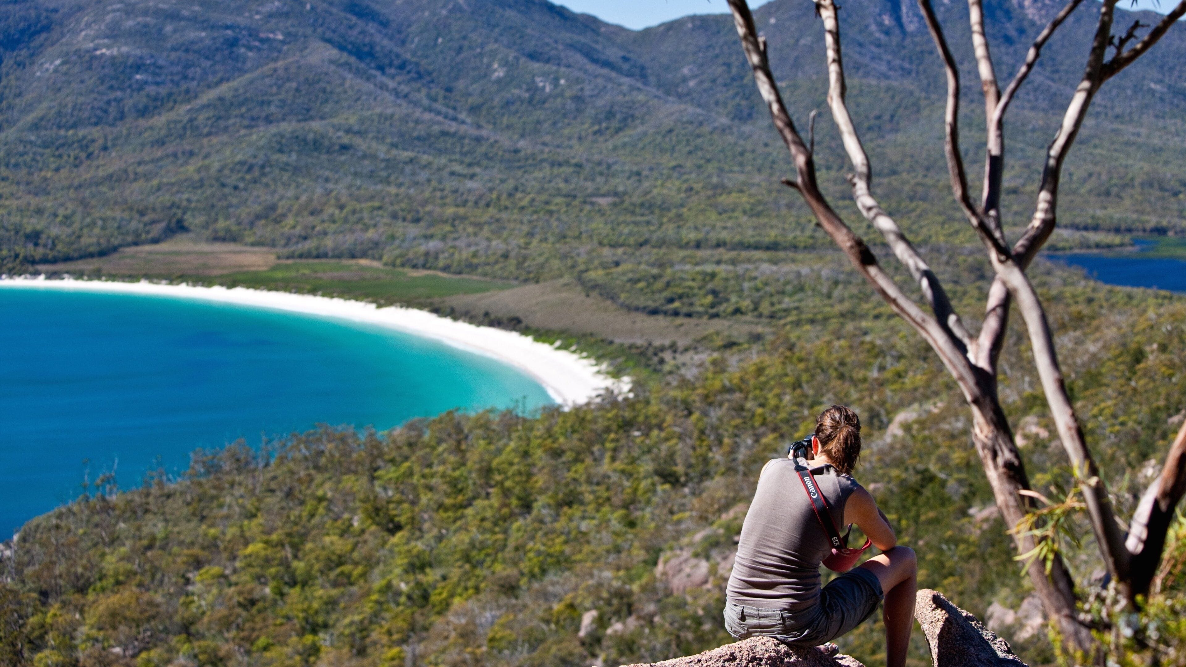 A person sitting on a rock overlooking a beach with clear blue water and green hills in the background.