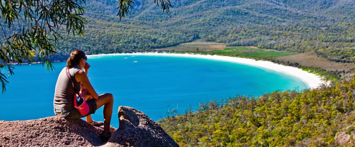 A woman sitting on a rock overlooking a turquoise bay with white sand beaches and green hills in the background.