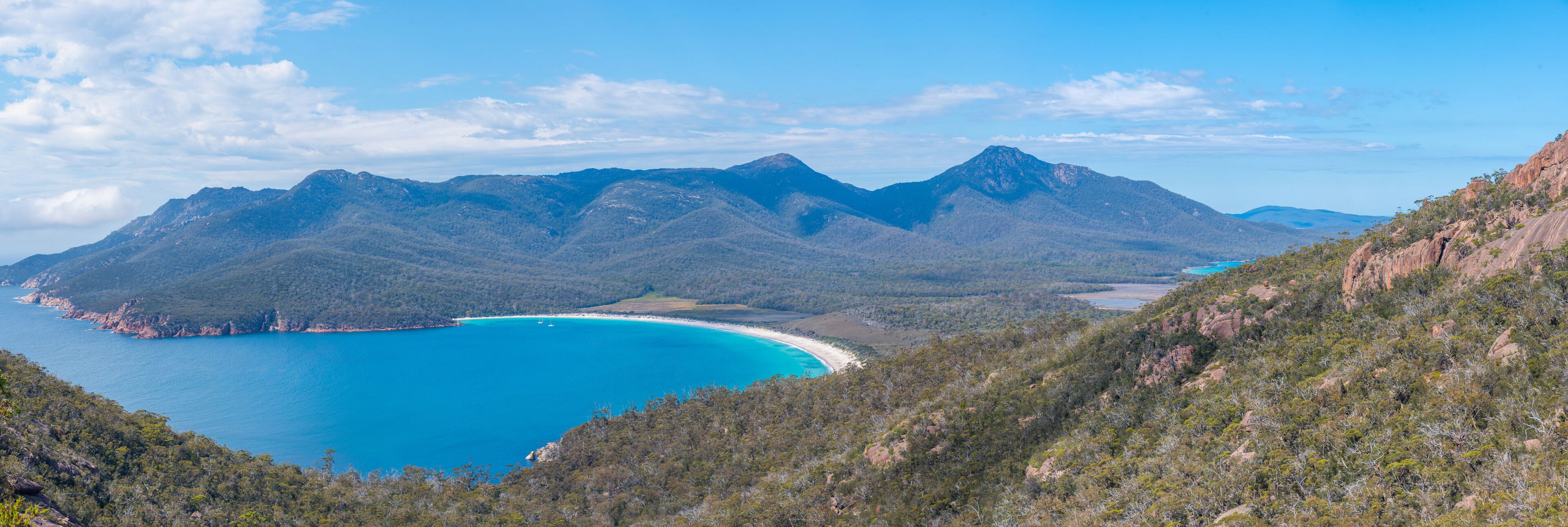 A coastal landscape with a turquoise bay, surrounded by green hills and mountains.