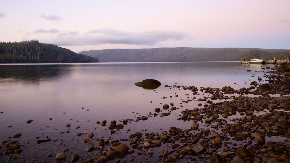 A calm lake with a rocky shoreline, a boat docked on the right, and a forested hill in the background.