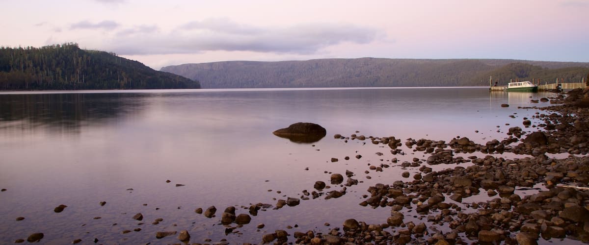 Tyyni järvi, jonka rannalla on kivikkoista maastoa, oikealla puolella laiturissa oleva vene ja taustalla metsäinen kukkula.