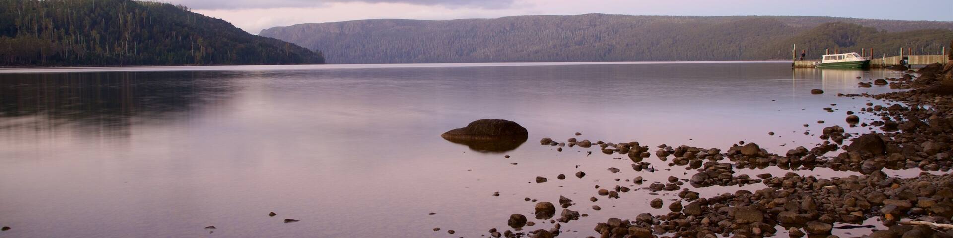 A calm lake with a rocky shoreline, a boat docked on the right, and a forested hill in the background.