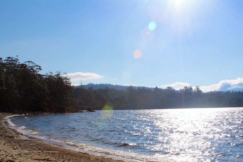 A beach with clear water, a sandy shore, and a forested hill in the background.